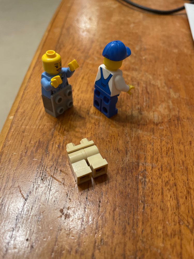 Two Lego minifigs.  The one on the left has their legs replaced with an arrangement presenting four studs forwards.  The one on the right is facing away from the camera demonstrating the stud holes in their rear. A pair of legs lies discarded on the table. 