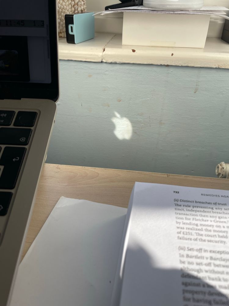 An reversed apple logo (of an apple shape with a bite out of it, with a leaf leaning in the direction of the bite) cast  on a blue wall behind a desk on which are a laptop and a trusts law textbook 