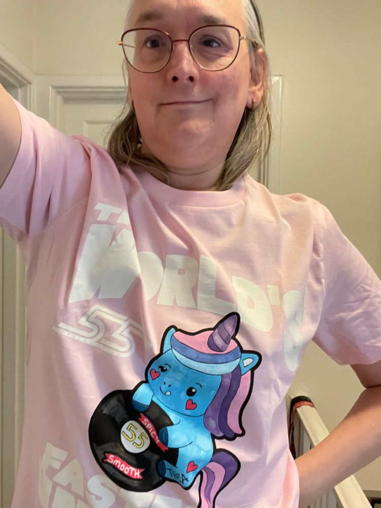 Girl in pink T-shirt. The T-shirt is labelled “The World’s Fastest Unicorn” with the number 55 and a drawing of a unicorn. The unicorn is blue with pink and purple hair and horn. She has hearts on her cheeks and her tongue is stuck out in close attention. She is holding a black tyre labelled “spicy” and “smooth” with the number 55 in the middle. She is also marked with Thea, her artist’s name. 