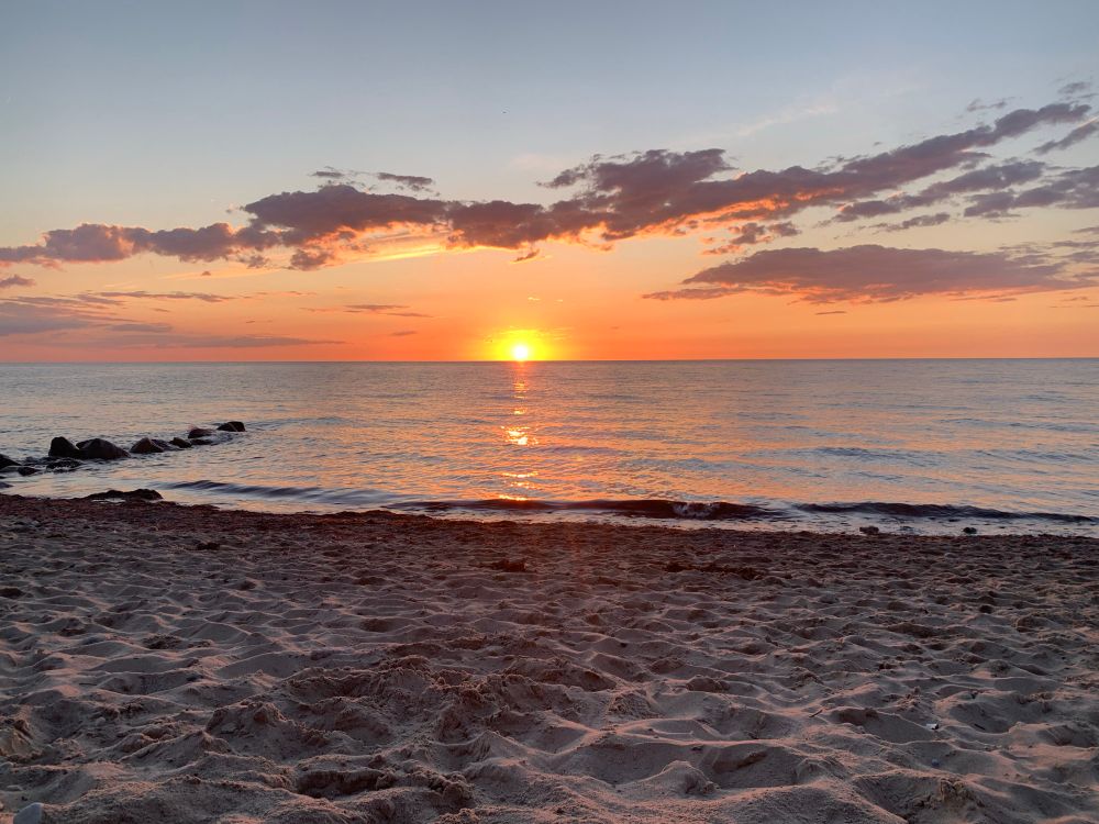Ostsee. Halbe orangene Sonne am Horizont. Einige Wolken, vorne Strand.
