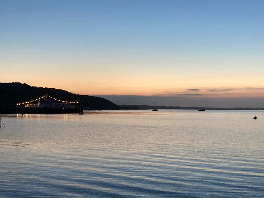 Ein mit Lichterketten geschmücktes kleines Passagierschiff auf der Förde, dahinter Wald, rechts davon einige vor Anker liegende Segelschiffe. Abendhimmel, keine Wolken, glattes Wasser.