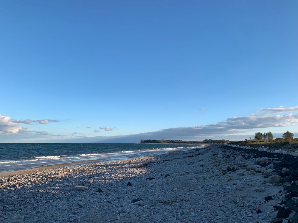Steiniger Steand an der Ostsee, links das Meer, im Hintergrund eine Landzunge mit Bäumen. Fast blauer hoher Himmel nur am Horizont einige Wolken.