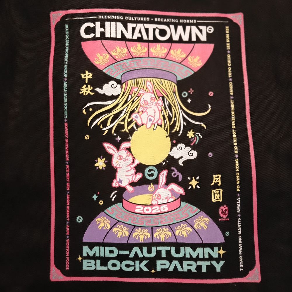 Close-up of the event tote. The tote is in black with an illustration of two chinese lanterns on the top and bottom and three bunnies coming out of it. There is a moon in the center with confetti-like details around it. 
Top text says "Chinatown"
Bottom text says "Mid-Autum Block Party"
The two main Chinese Text says "Autumn" and "Full Moon" 