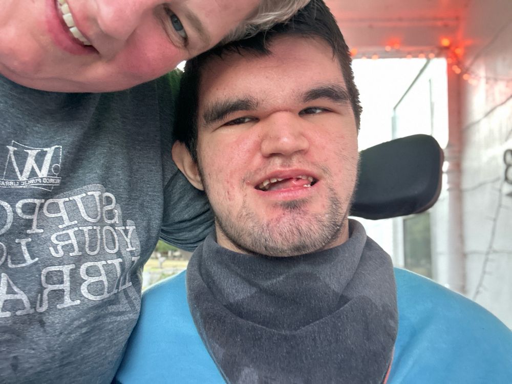 Picture of a young man with dark hair and brown eyes, smiling. He is wearing a blue shirt and a gray bandana. He is sitting in a wheelchair. A woman has her arm around his back and is leaning over. She is wearing a “support your local library” t-shirt. 