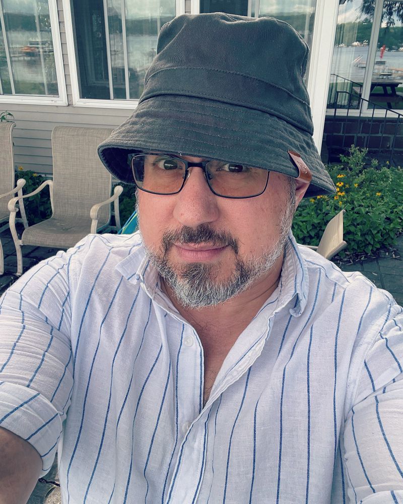 A bearded white man in glasses, a white button down shirt with blue stripes, wearing a green canvas bucket hat. It is ridiculous. 