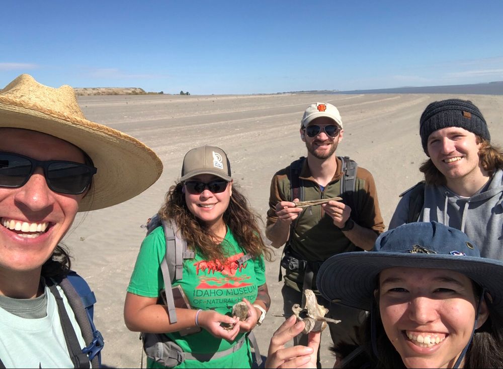 Group selfie on an Idaho beach: wide sandy expanse and blue skies. Sloth vertebra in the foreground!