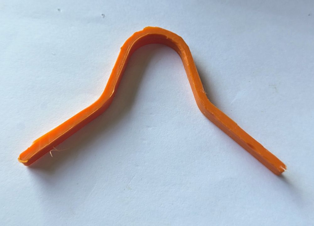 Thin orange 3d printed plastic strip in shape of a mask's nose bridge on a white paper sheet. It's got an few rough edges along the top side that need sanding. 