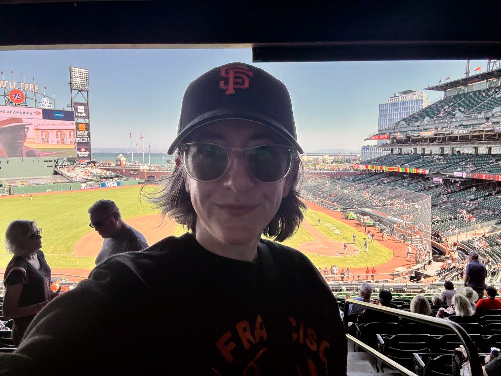 Person in San Francisco Giants gear takes a selfie inside Oracle Park with the field and scoreboard in the background.