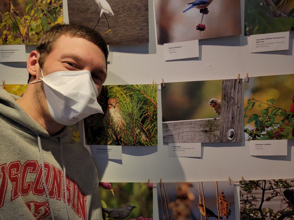 photo of a man wearing an N95 mask standing in front of a wall of photos featuring birds. he's next to one with a pine siskin peeking out around a fence post.