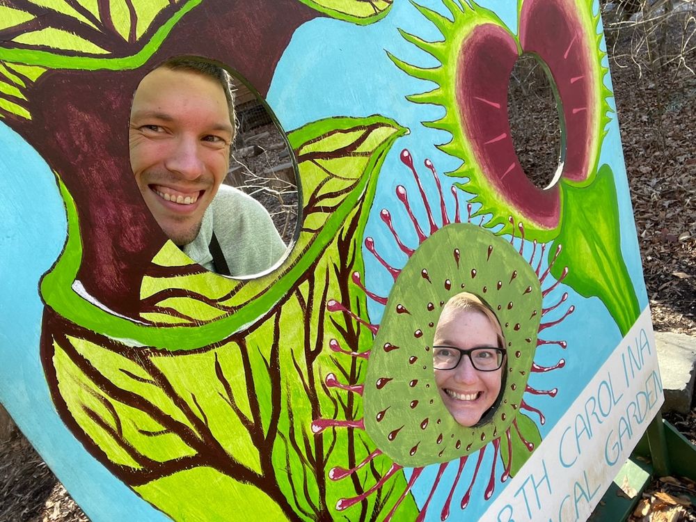 selfie of two people on a carnivorous plant face cutout. the guy is a pitcher plant and the femme is a sundew. the Venus flytrap is sadly empty.