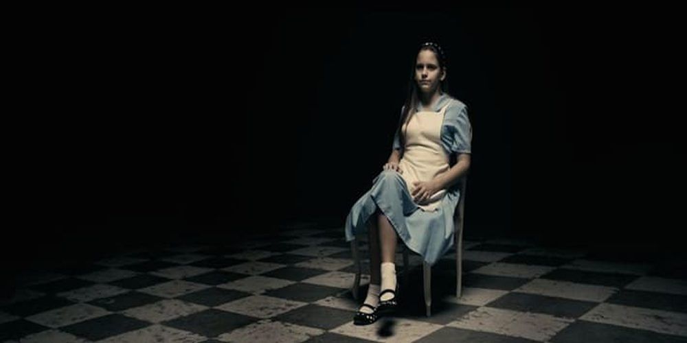 Photogramme de A Serbian Film.
Un personnage féminin vêtu d'une robe bleu et chaussé de chausettes blanches et de ballerines noires est assis sur une chaise, jambes croisées, dans une pièce carrelée de noir et de blanc. Elle regarde quelque chose hors champ (je te dis pas quoi parce que la censure ma caille mais si tu sais tu sais).