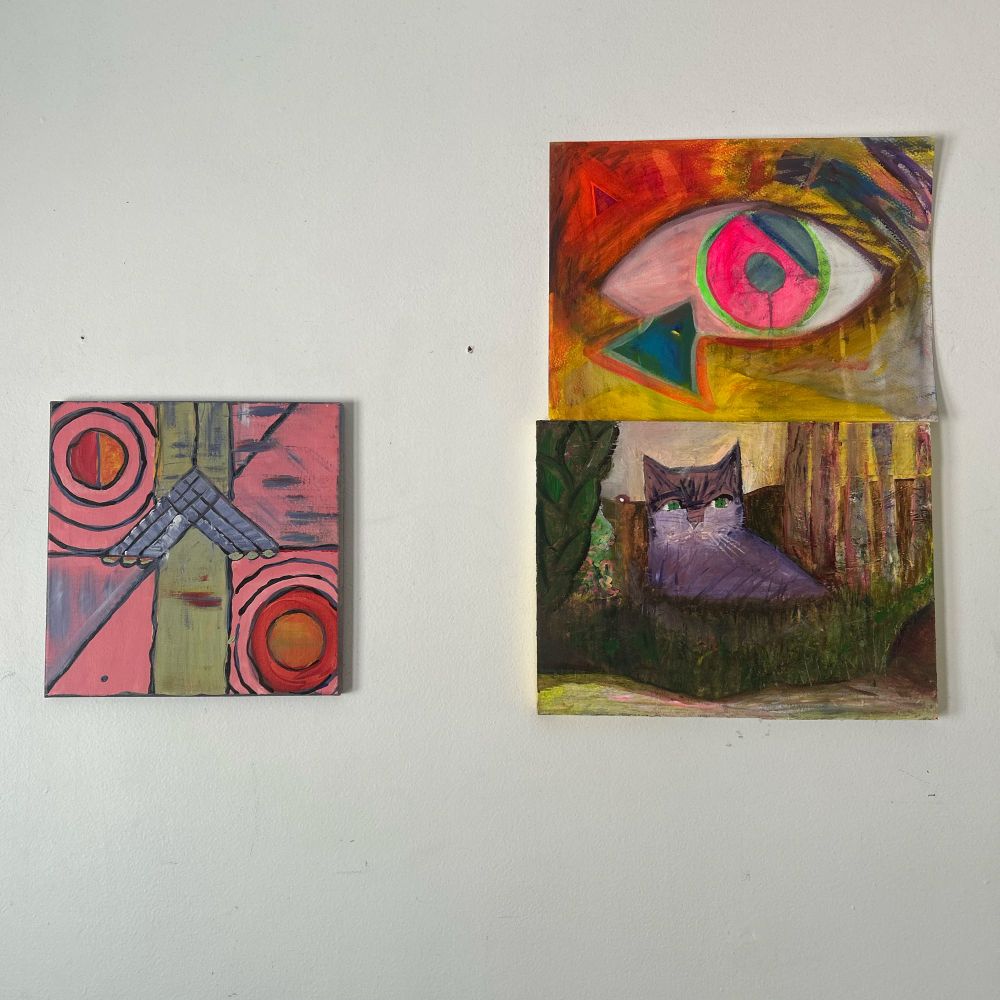 A picture of a painting of an arrow pointing up, symbolizing forward movement. It is surrounded by two diagonal suns shining, and has a line running through the other diagonal. The background is pink. To the right are two paintings stacked on top of each other; the top one is a drippy, trippy, luminous painting of an eye signifying consciousness, and the one below is a painting of a purple tabby cat in a farm field surrounded by brush and trees with a red barn with a light on inside in the distance.