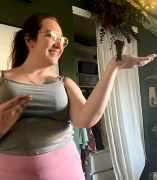 a photo of me, a white woman with brown hair in a pony tale. i'm smiling widely and holding my hand up like the "is this a pigeon?" meme. nuggie, a male common house sparrow, is perched on my hand, head cocked towards me.