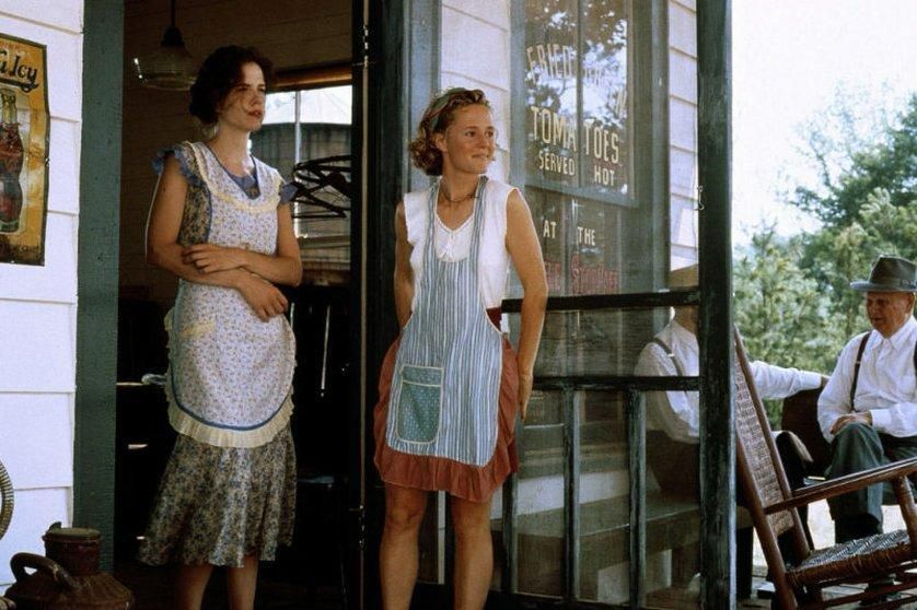 Dos mujeres de pie con delantal mirando hacia el exterior en el porche de un establecimiento de comidas en los años 30. Es un fotograma de la película tomates verdes fritos y ellas son las protagonistas