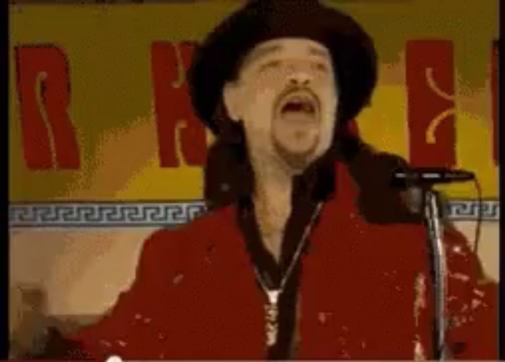 a man is singing into a microphone while wearing a red jacket and hat .