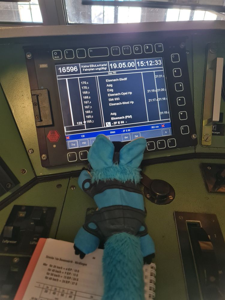 Protobean plushie lying in front of a digital driver's timetable in German trains with software from around the year 2000