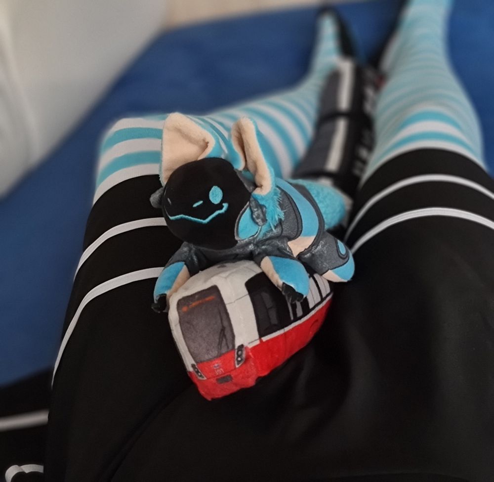 Protobean plush on top of a Wiener Linien Flexity tram plush lying inbetween a femboy's legs with blue-white striped thigh highs and a black skirt with two white stripes