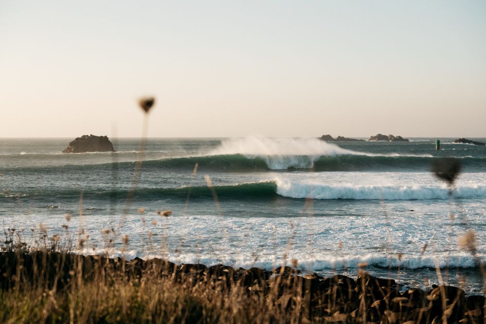 A scenic view of ocean waves crashing against the shore, with rocky formations in the background. Tall grass is slightly in the foreground, adding depth to the image. The atmosphere is tranquil, with a soft light from the setting or rising sun.