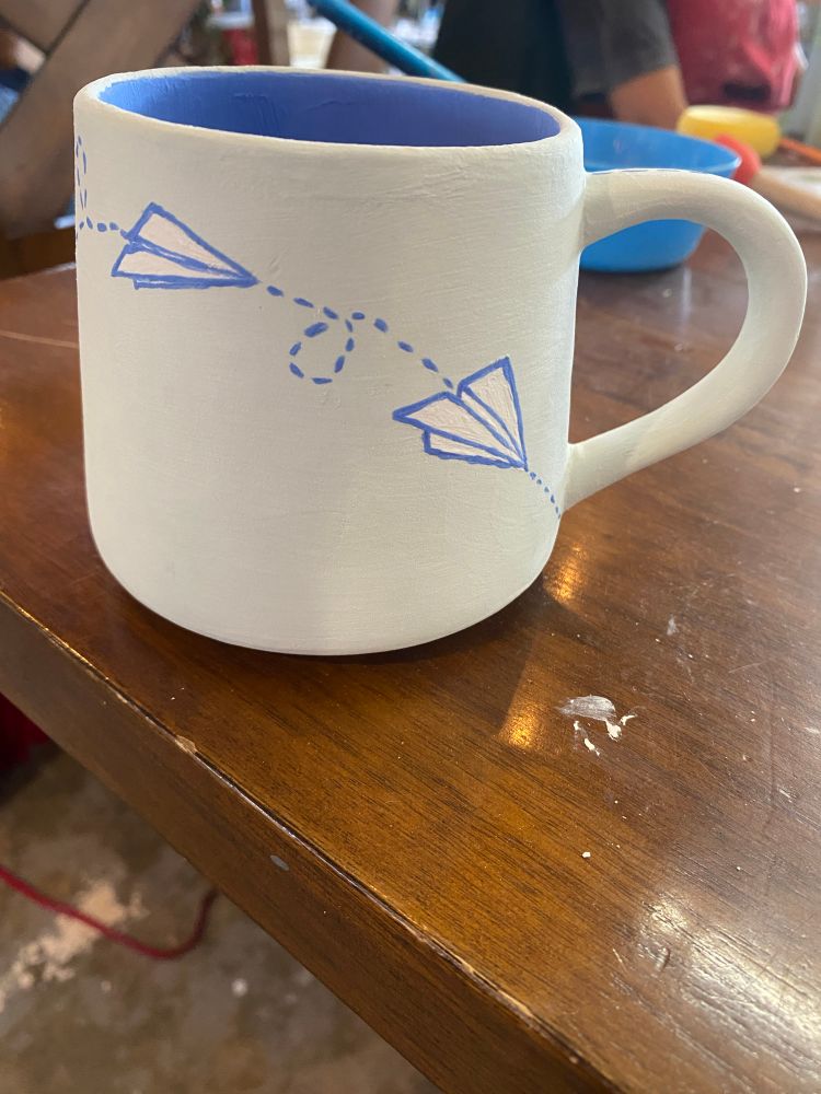 Other side of the ceramic mug painted with a paper airplane flying across 