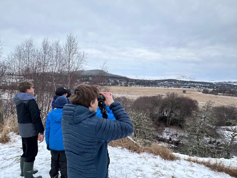 Three individuals using binoculars observe a wintry landscape with distant hills under a cloudy sky.