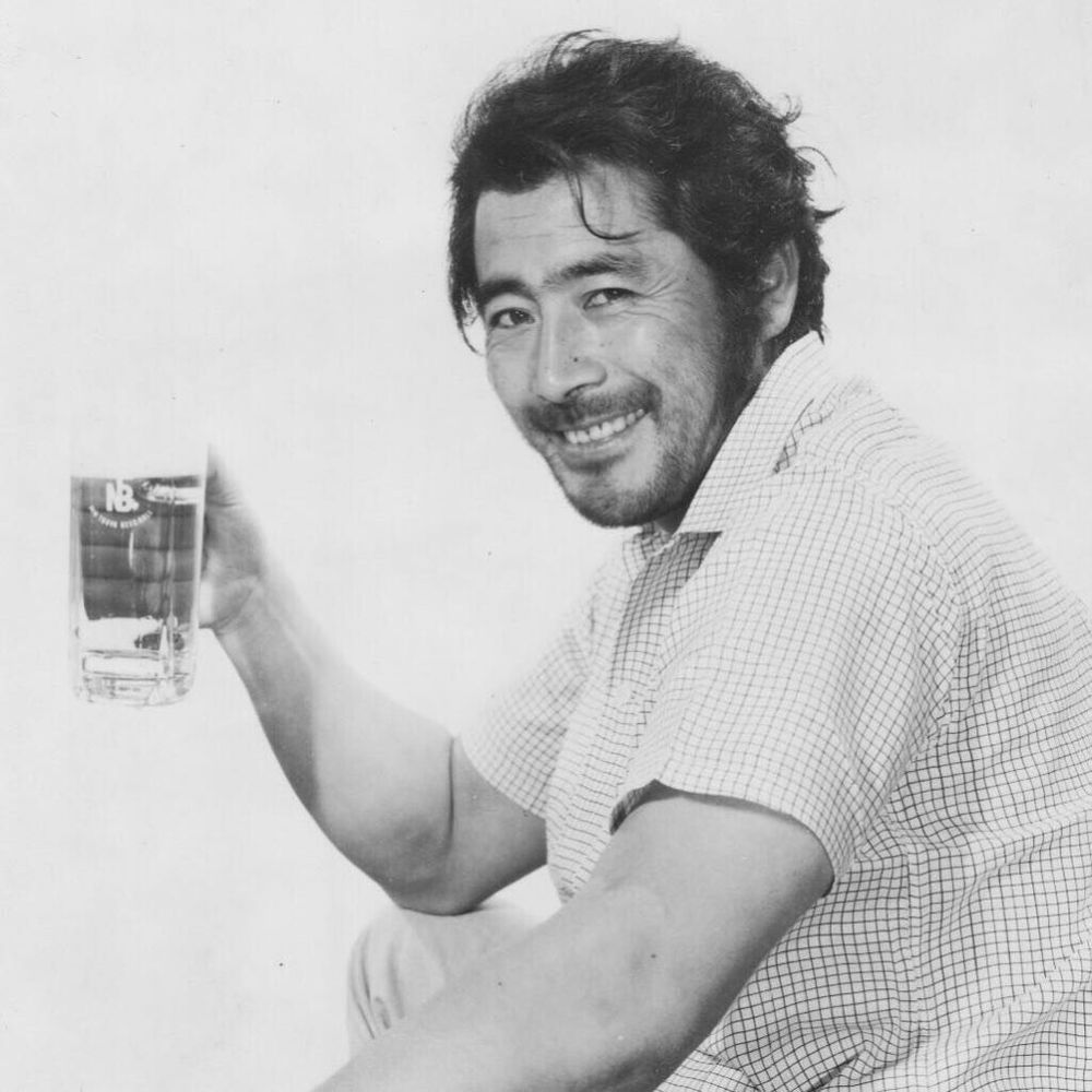 Absurdly handsome Japanese actor Toshiro Mifune smiling at the camera and holding up a drink.