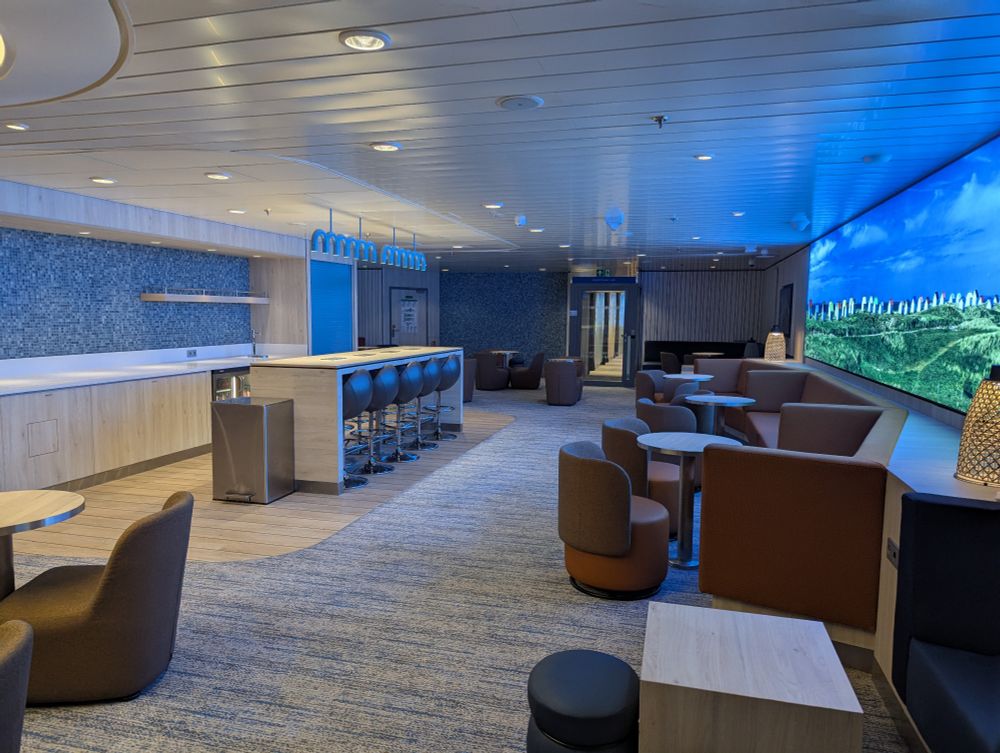 Lounge area on Guillaume de Normandie for Couchettes Plus passengers exclusively. Bar area with water dispenser on left, comfortable seating against illuminated photo of beach huts on right.