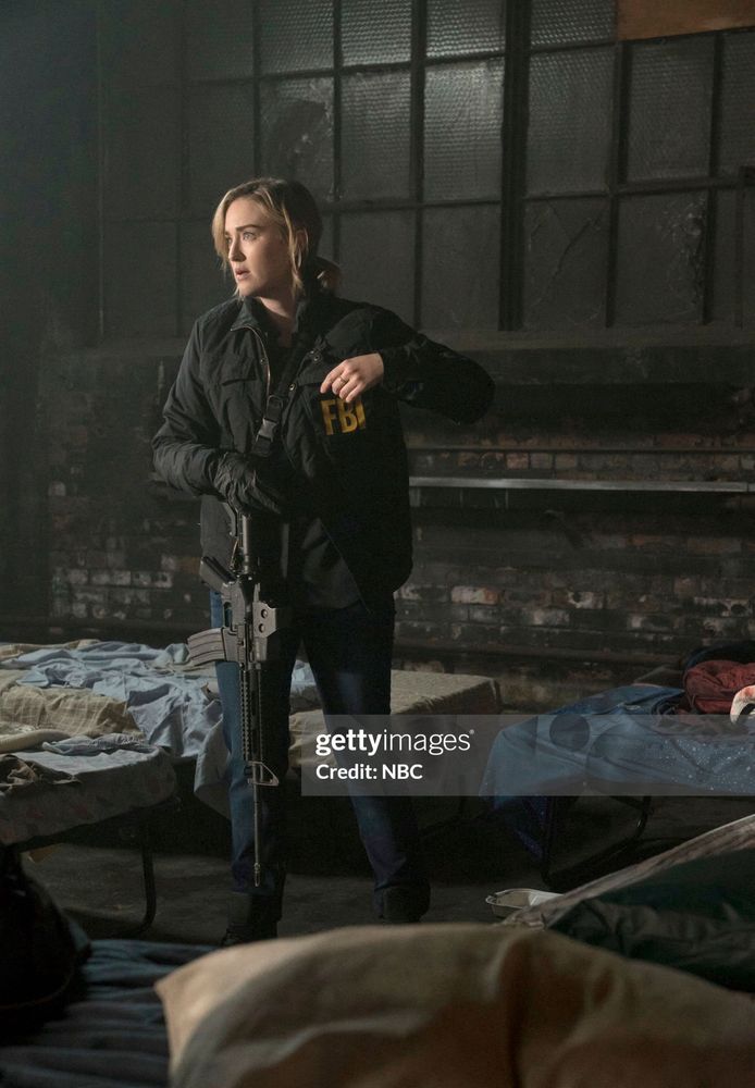 A Getty images photo from NBC’s Blindspot of Ashley Johnson portraying Patterson. She is wearing jeans, a coat that has “FBI” on the front of it, and has a large gun strapped across her body, hanging down by her side. She is standing amongst what looks like some cots that are set up in a warehouse. Her face looks concerned and a little surprised, and she is looking off to her right