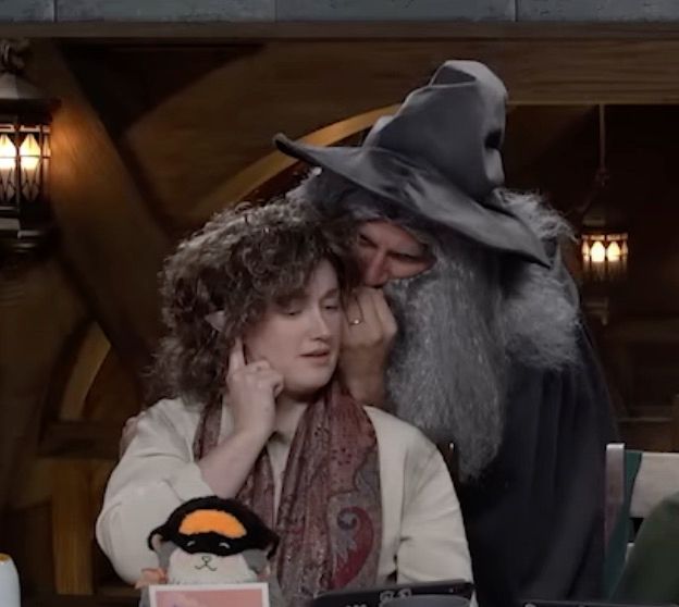 Ashley Johnson dressed as Pippin or Merry from Lord of the Rings. She is sitting at the Critical Role table with a short curly wig, elf ears, a peasant tunic like shirt, and a scarf. She has her right hand plugging her right ear, and standing behind her is Matt Mercer dressed as Gandolf whispering into her left ear.