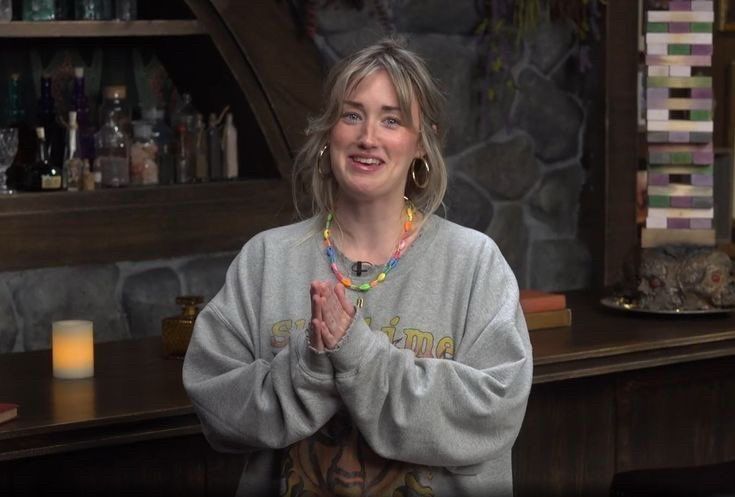 Ashley Johnson hosting 4 Sided Dive. She is standing in front of the tavern’s bar doing the intro to the show wearing a gray crewneck sweatshirt that says “sublime” with an art piece that’s covered by her hands touching in front of her. Her hair is pulled back and she is wearing a colorful necklace. She is smiling at the camera.