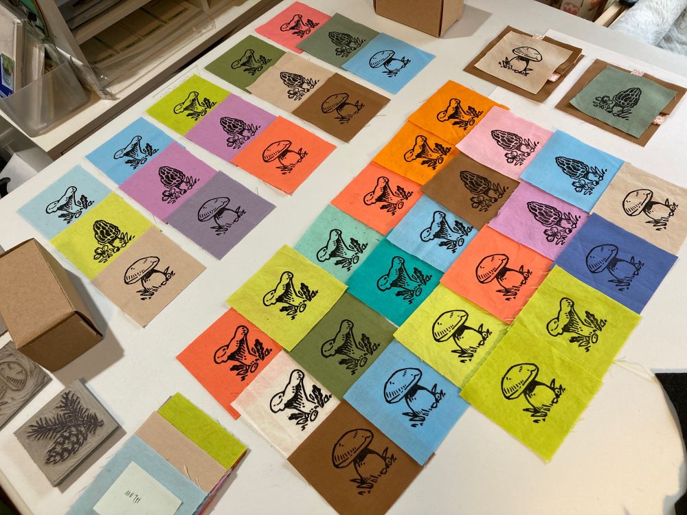 a tabletop spread of many linocut prints of mushrooms on many bright colored squares of fabric 