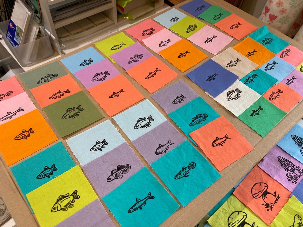a tabletop spread of many linocut prints of fish on tiny squares of brightly colored fabric 