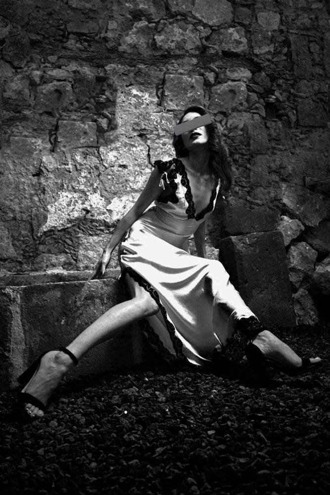 Black and white photo of a woman in a silk dress posing while sitting down with legs outreached chest leaned forward. Backdrop is a cobblestone wall. 
