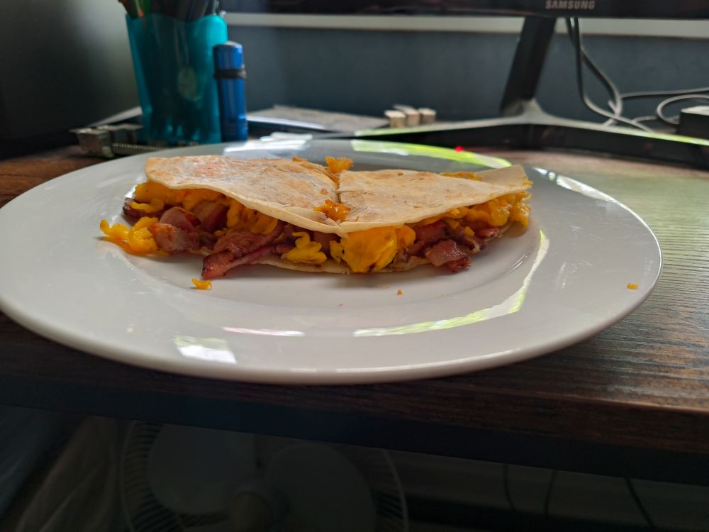 A breakfast quesadilla, with bacon and egg, on a white plate. it is sitting on a desk with a fan underneath and a pot of pens behind and off to the left of the plate