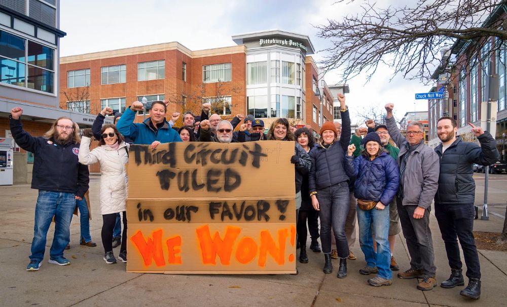 A group of people holding up their fists behind a sign that reads "Third Circuit ruled in our favor! We won!"