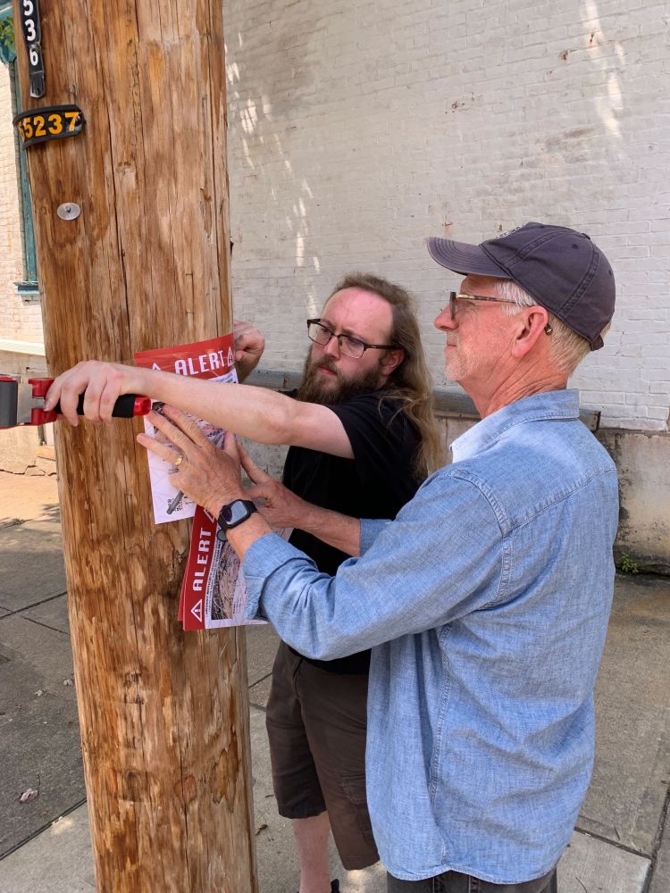 Two workers tape a white sign with a red heading to a brown utility pole.
