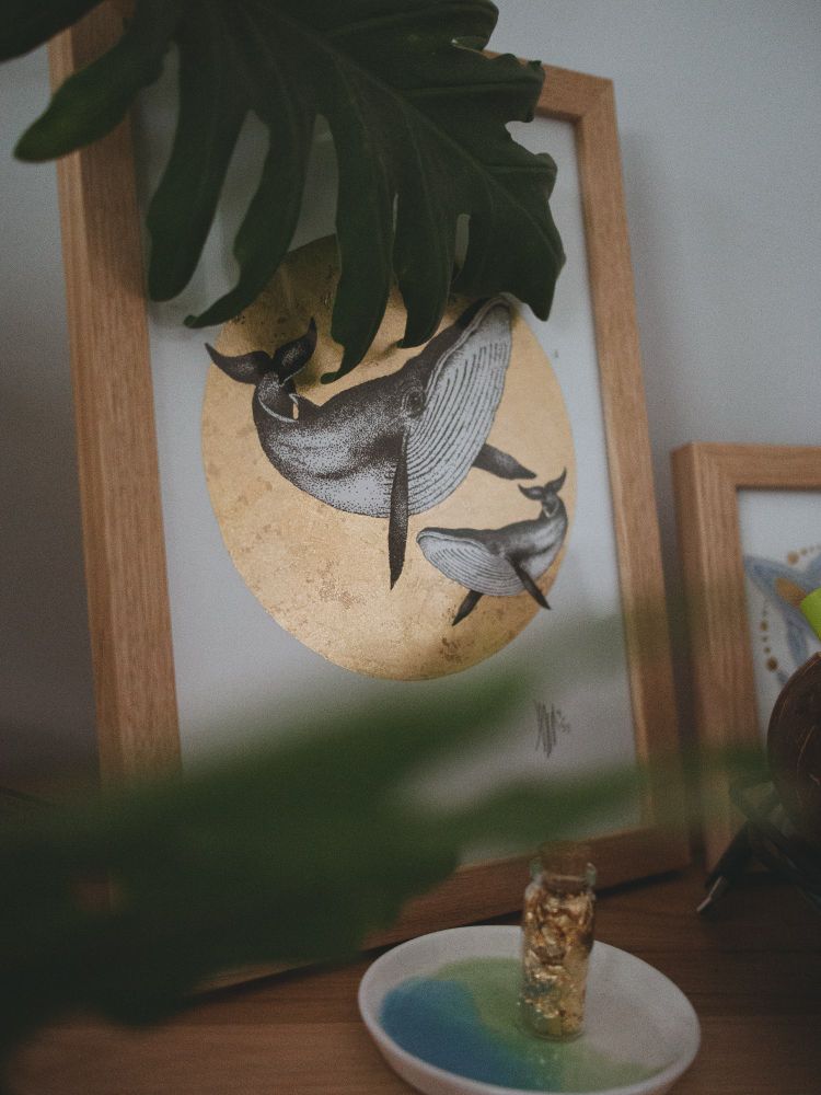 A photo of a piece of art by apothecaryartist framed in a light oak frame. Two whales are drawn in dotted ink, sitting in a circle of gold leaf.
Some monstera leaves partially obscure the frame.