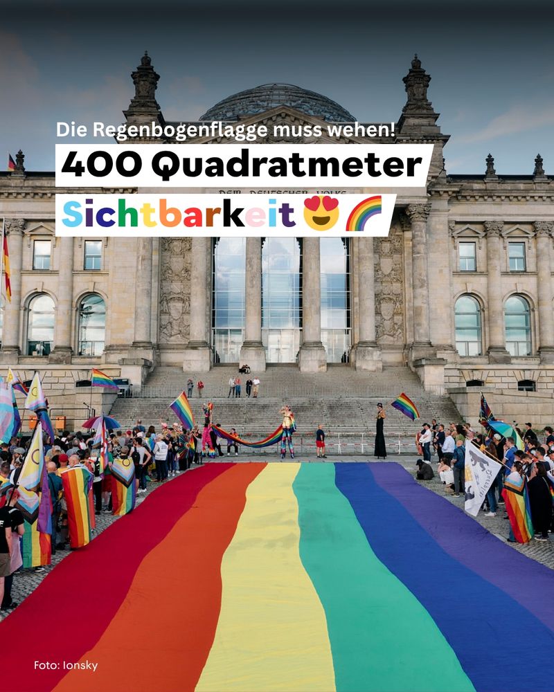 Menschen stehen dicht gedrängt an einer bunten Stoffbahn, viele halten oder schwenken Regenbogenflaggen. Die Stimmung wirkt ausgelassen und feierlich vor dem Berliner Reichstag.