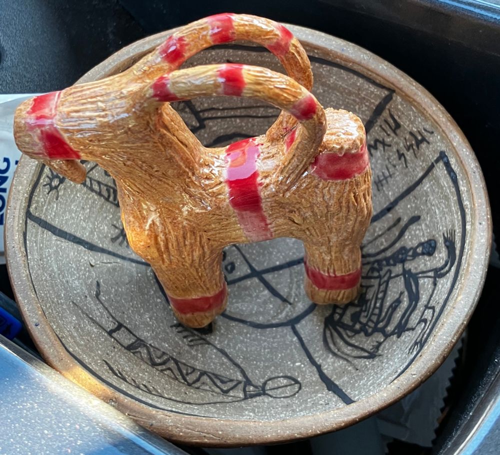 Small clay goat, now fired with red glaze for the ribbon bits & a straw-color glaze for the body, stands in an incantation bowl tucked into a car cubby for transport home from the clay studio.