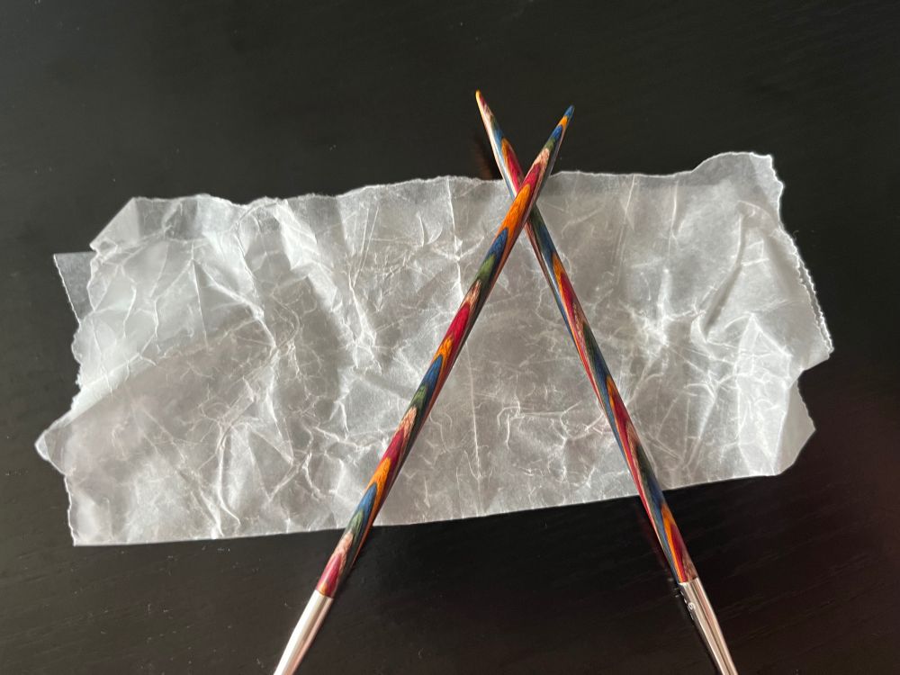 Two multi-coloured Knit Picks brand circular knitting needles crossed on a piece of rumpled wax paper with a black background.
