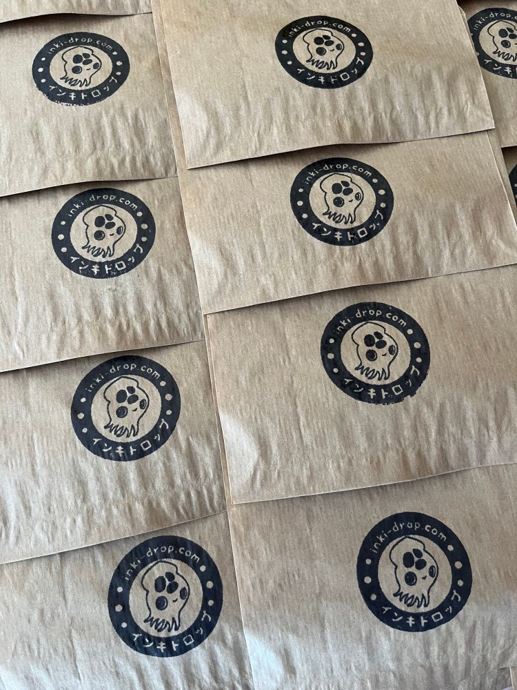 Photo of brown paper bags stamped with a black logo. Logo features a squid encircled by text in english and japanese.  Text reads “inki-drop.com” 