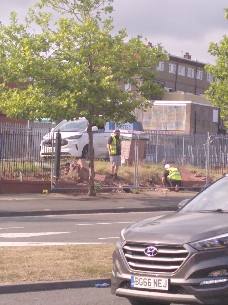 2 men in hi-vis tops working on a fallen section of wall behind a tall, wire fencing.