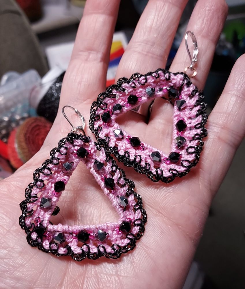 My left hand holding a pair of earrings created out of copper wire then micro crocheted in pink cotton thread with details in metallic black thread and beaded with black and silver conical beads and hot pink seed beads.Silver French leverback findings.
