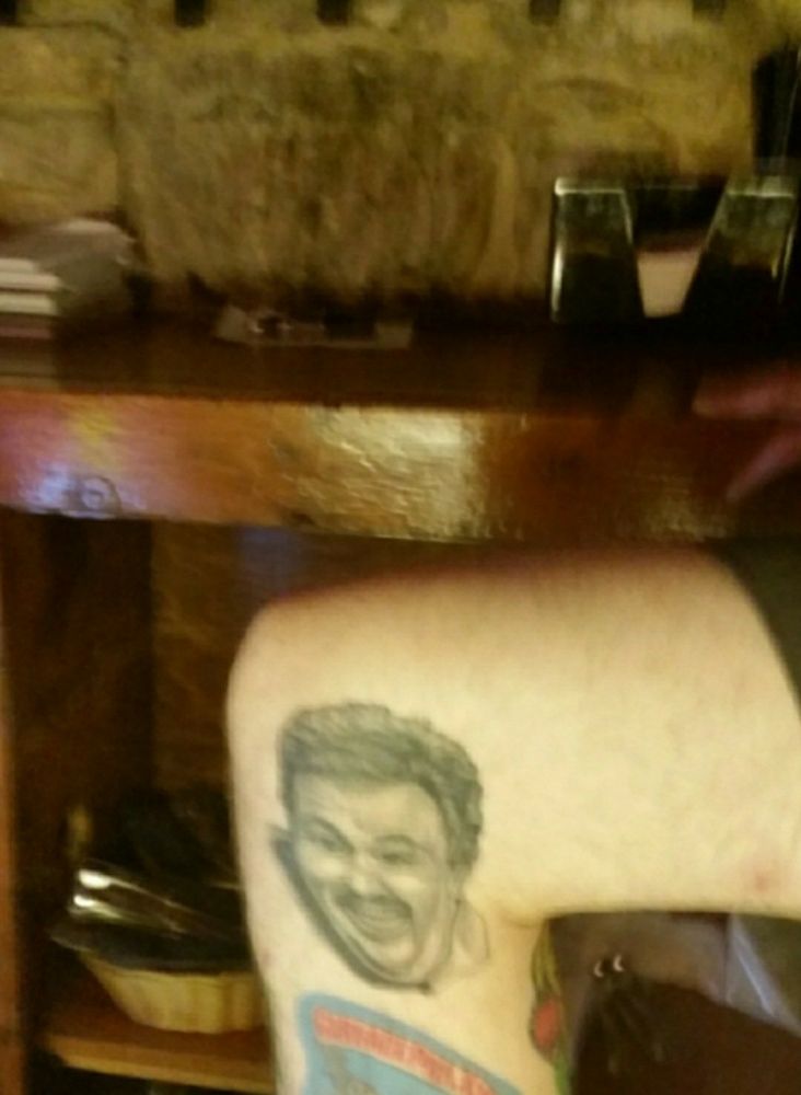 Photo of a Maryland bartender's right thigh and leg. The left side of his right calf is tattooed with the black and white likeness of John Candy as Del Griffith.  Not fully pictured: a colorful tattoo of the Garbage Pail Kids is directly under the John Candy tattoo. 