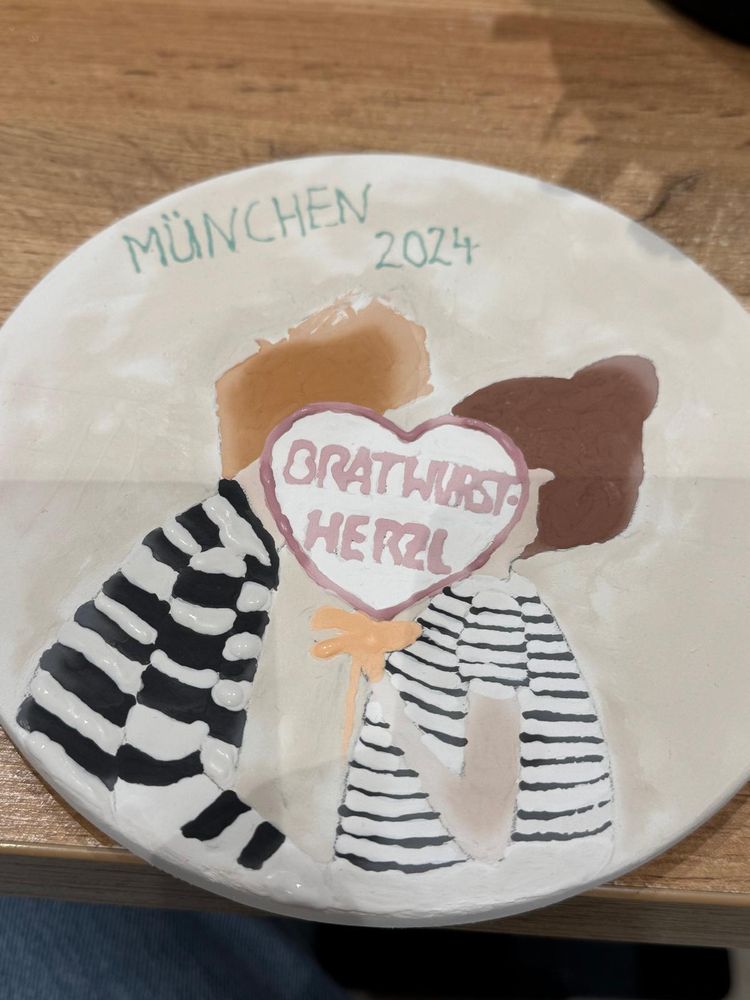 A plate that shows two people kissing behind a heartshaped sign that reads "Bratwurstherzl" ("grilled sausage heart") and "Munich 2024"