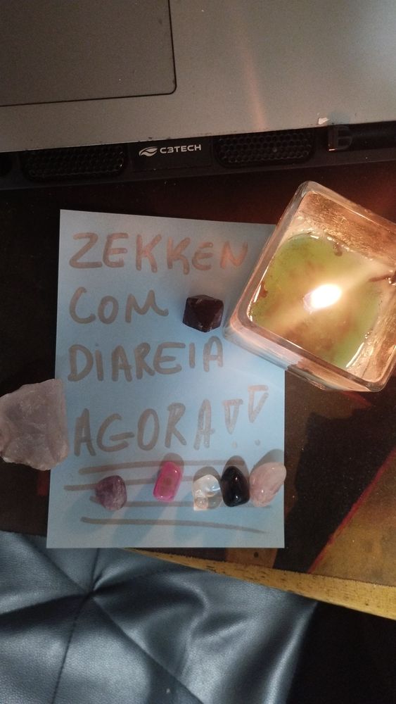 Foto de um papel ao lado de uma vela acessa com varias pedras em cima. No papel se le, em letras maiúsculas"ZEKKEN COM DIARREIA AGORA"