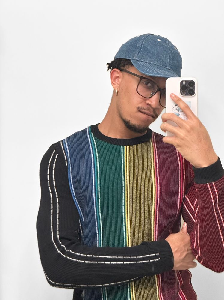 fuhtrue taking a bathroom mirror selfie. he’s wearing a denim cap, obnoxiously big box-frame glasses, silver hoop earrings, and a ribbed rainbow sweater.
