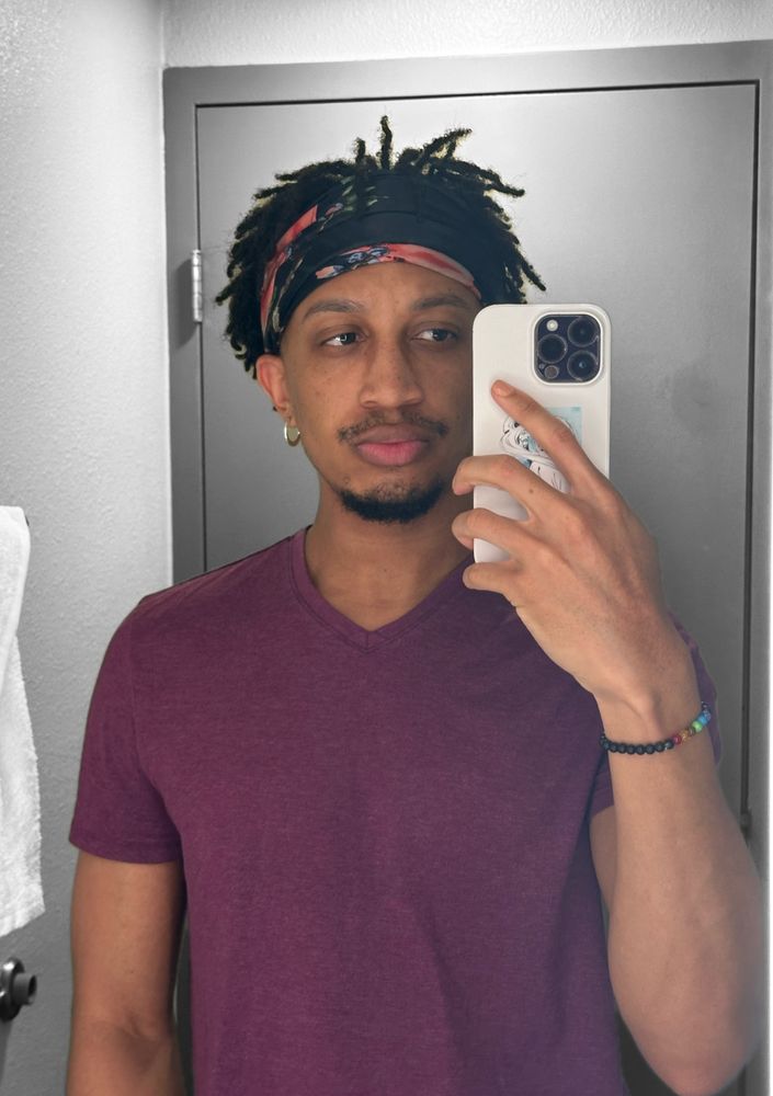 fuhtrue standing in front of a hotel bathroom mirror taking a selfie. he’s wearing a scarf to hold his locs out his face, silver hoop earrings, a chakra bracelet, and a purple v-neck.