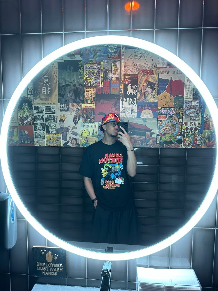 fuhtrue once again in bathroom taking a mirror selfie. he’s wearing a tie-dye rainbow bucket hat with a distorted yin yang sign, a chakra bracelet, an apple watch, a Levar Burton graphic tee, black adidas cargo pants, an apple watch and his round glasses.