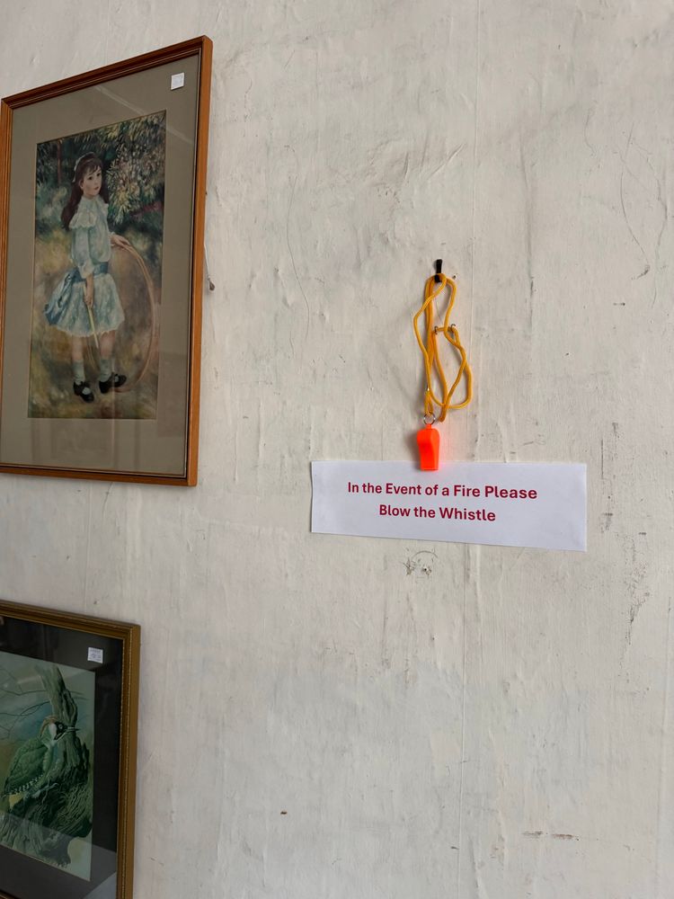 In the middle of a wall, and orange whistle on a yellow string hangs from a hook. A sign beneath it reads “In the Event of a Fire Please Blow the Whistle”.