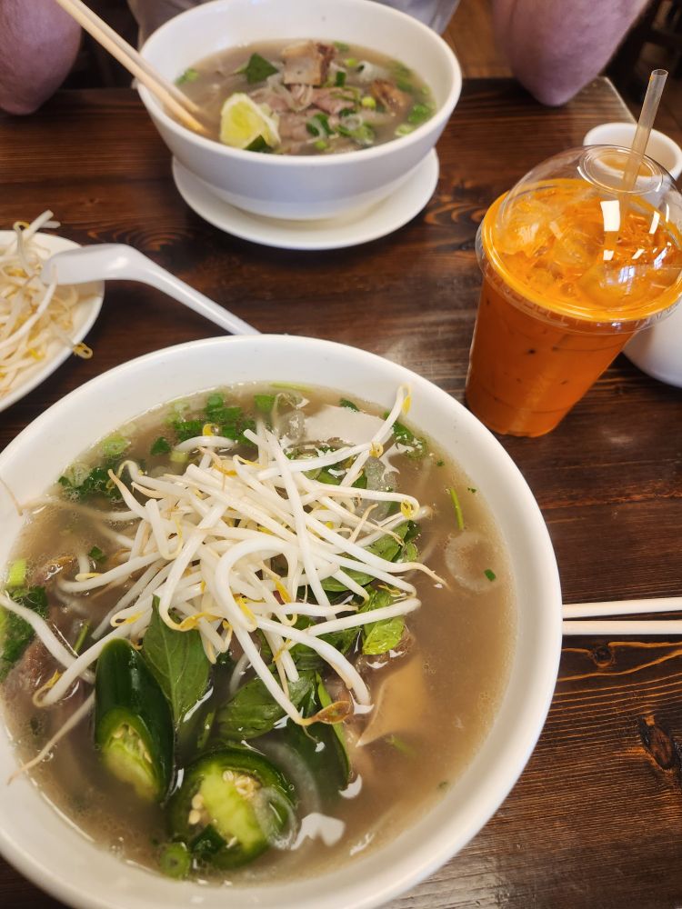 A delicious bowl of pho, in the background a friends delicious bowl of pho. It is accompanied by a thai iced tea. 
